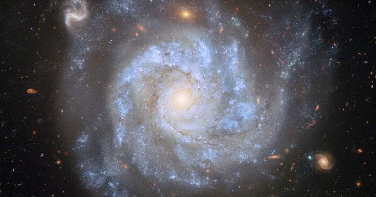 A top-down view of a spiral galaxy, showing its brightly shining centre, its broad spiral arms and the faint halo around its disc, as well as distant galaxies and stars on a dark background (Cover Image Source: ESA Hubble)