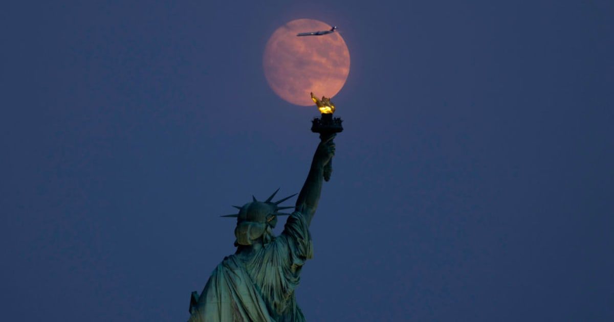 The Harvest Moon illuminated at 98 percent rises behind the Statue of Liberty in New York City as an airplane flies past one day before turning full on September 16, 2024 (Cover Image Source: Getty | Gary Hershorn)
