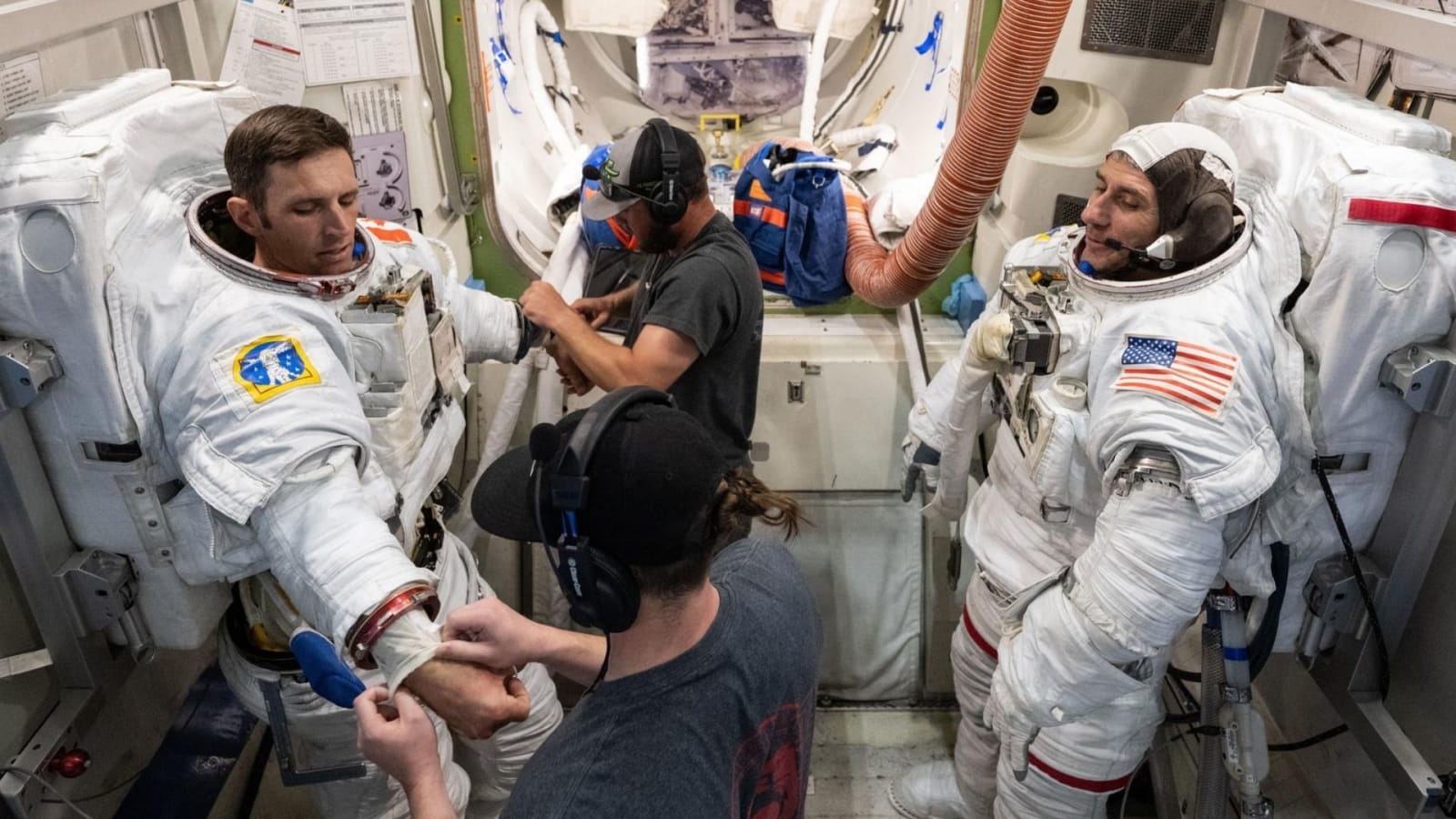 Josh Kutryk (left) and Luke Delaney (right), with assistance from training specialists, practice suiting up in spacesuits in an International Space Station airlock simulator at Johnson Space Center.